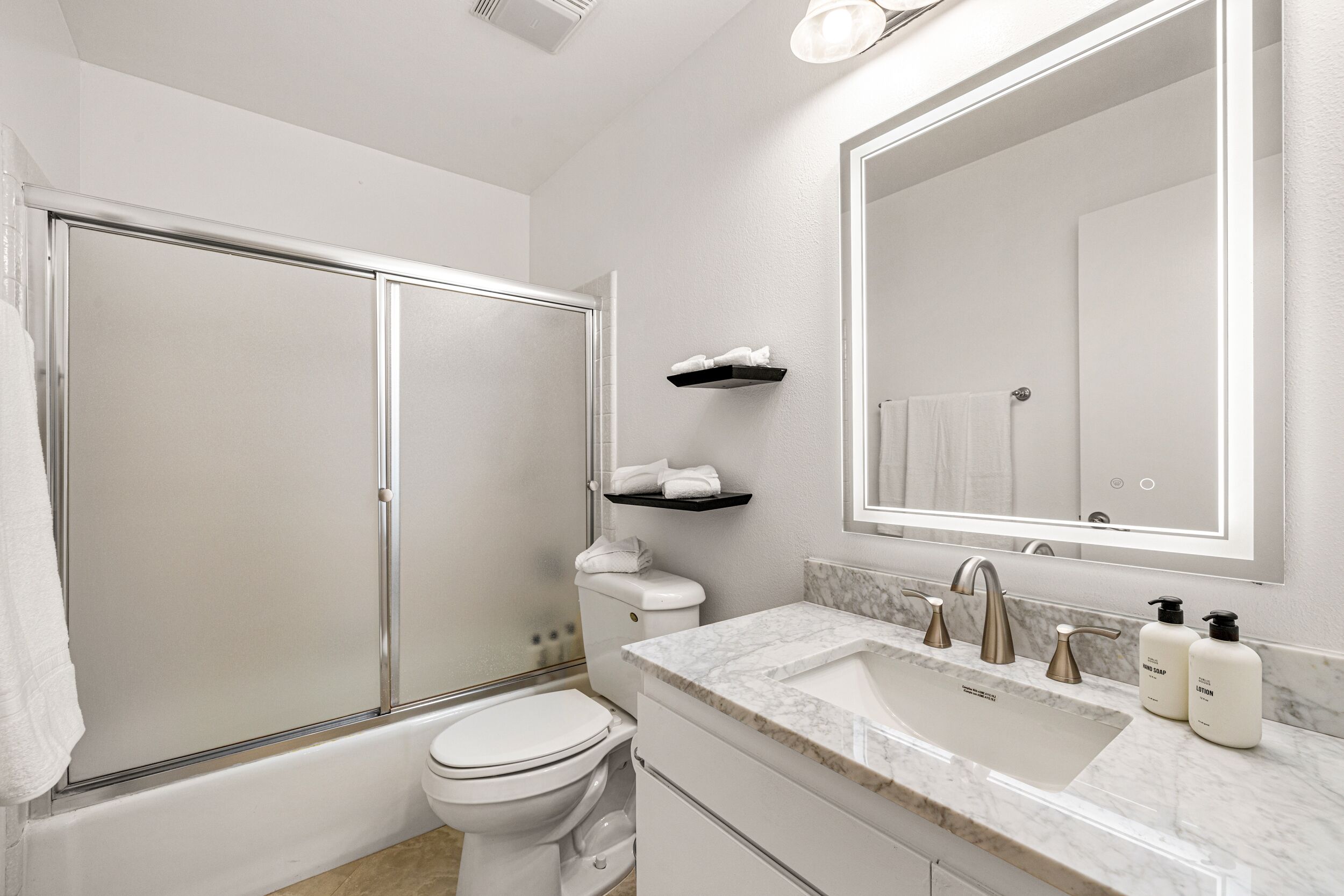 A modern bathroom with a glass-enclosed shower, a white toilet, a marble sink, and a large mirror.