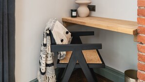 Un rincón de una habitación con una silla negra, un estante de madera, un jarrón con flores secas y un cuadro en la pared.