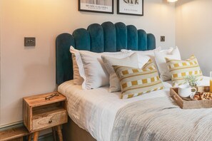 Un lit bien fait, avec une tête de lit recouverte d’un tissu turquoise, du linge de lit blanc et des coussins à motifs. Une table de chevet en bois, dotée d’un tiroir, et une paire de lunettes sont posées à côté du lit. Une assiette avec une tasse et des viennoiseries se trouve sur le lit.