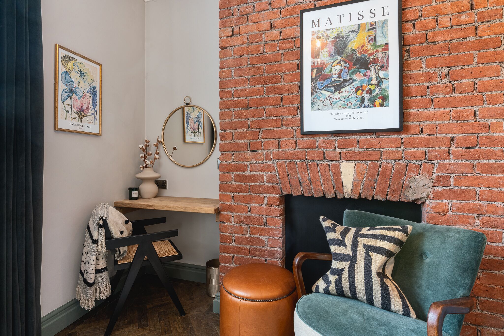 A room with a brick fireplace, a green armchair, a wooden desk, and a framed artwork of Matisse.