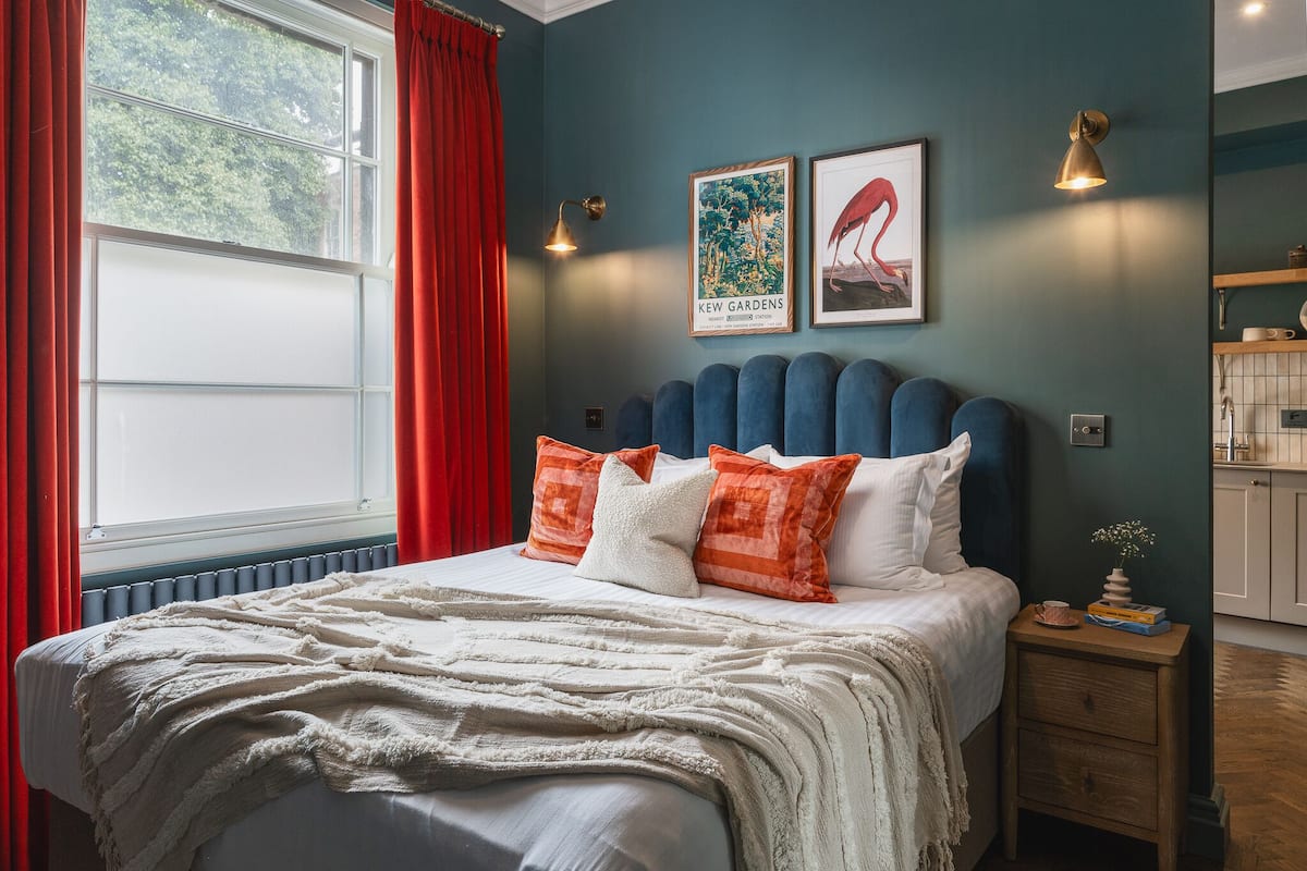 A bedroom with a bed, red curtains, a window, a bedside table, and a wall with two framed pictures.