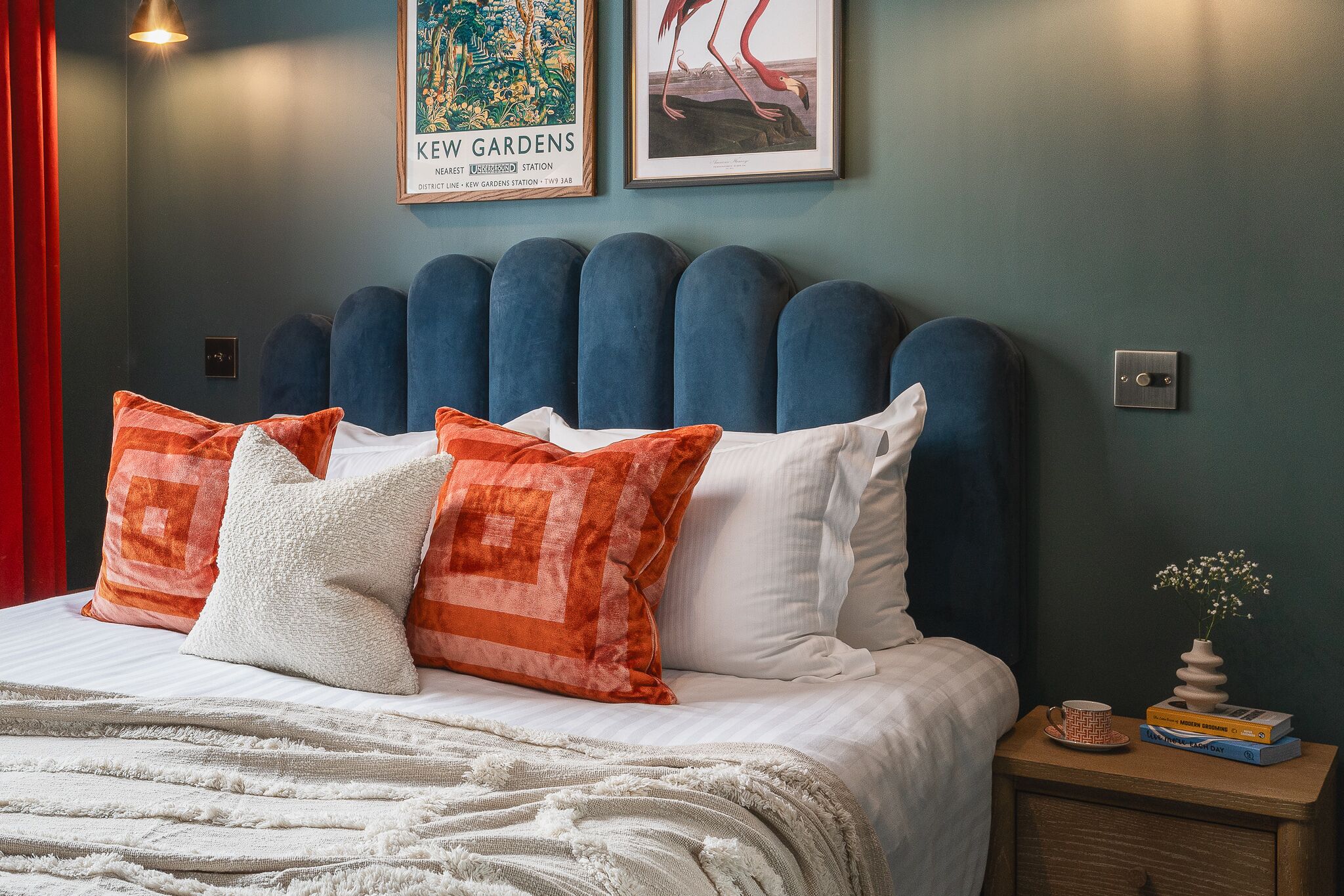 Un lit bien fait, avec une tête de lit rembourrée bleue, des coussins blancs et orange, un plaid blanc et une table de chevet ornée d’un vase et de livres.