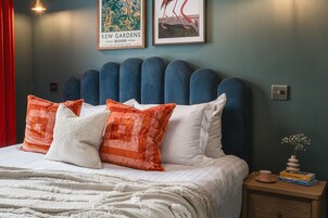 Una cama bien hecha con un cabecero tapizado en azul, almohadas blancas y anaranjadas, una manta blanca y una mesita de noche con un florero y libros.
