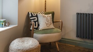 Un rincón acogedor con un sillón tapizado de verde, un reposapiés tejido, una lámpara y un radiador.