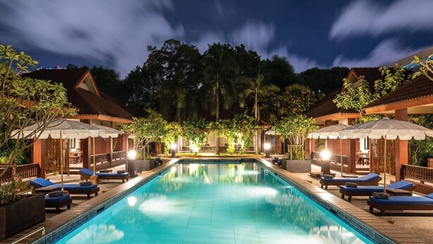 A swimming pool with lounge chairs and umbrellas, surrounded by buildings and trees. - RB Resort (Pattaya)