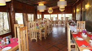 A spacious dining area with wooden tables and chairs, large windows, and a tiled floor.