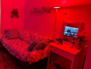 Quarto com cama iluminada em vermelho, uma mesinha com espelho e televisão na parede.