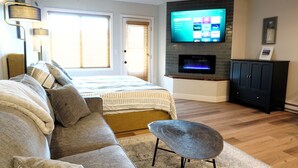 A living room with a sofa, a coffee table, a flat-screen TV mounted on a brick wall, and a fireplace below it. - GREAT Value! Classy Comfortable King Bed Studio, Sleeps 4! (Granby)