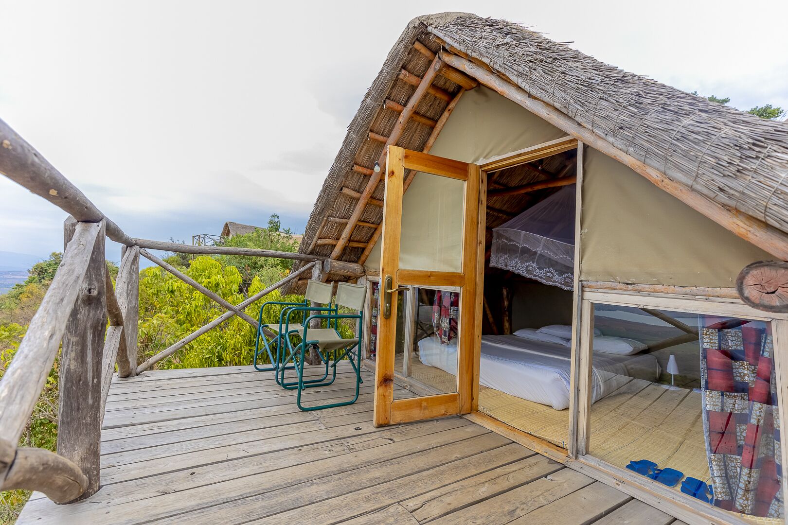 A thatched-roof tent with a wooden deck, a bed, and a chair.