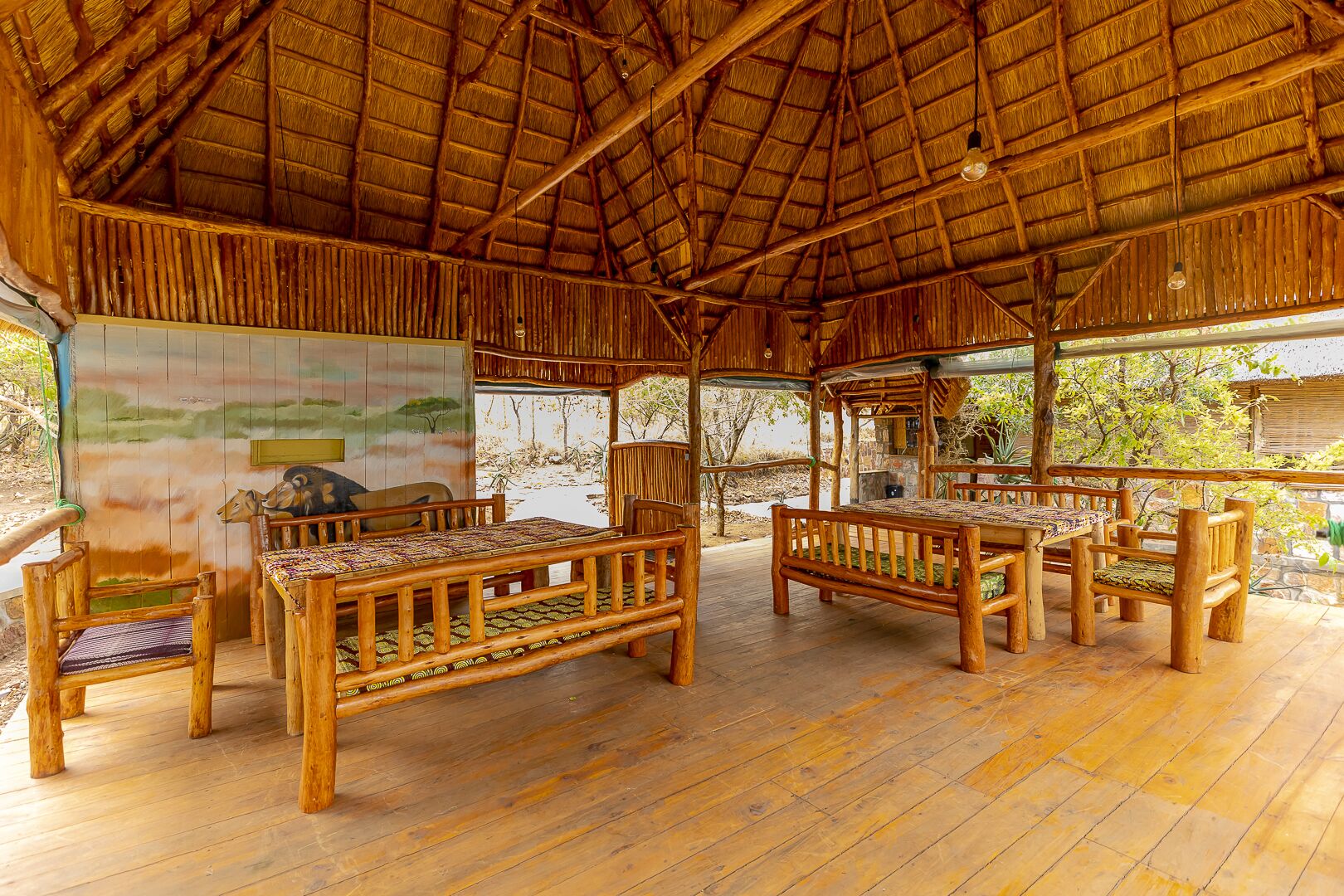 A rustic wooden hut with a thatched roof, furnished with wooden benches and a table, and a mural on the wall.