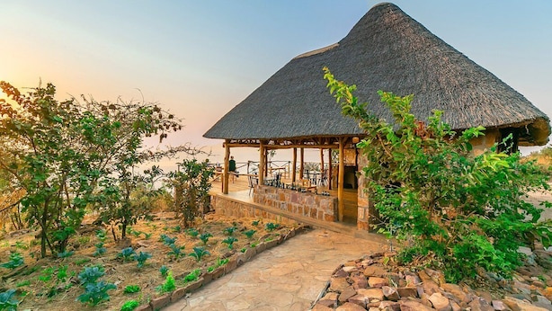 A thatched-roof pavilion with outdoor seating, surrounded by a garden with various plants and a stone pathway. - Akagera Rhino Lodge (Kabare)