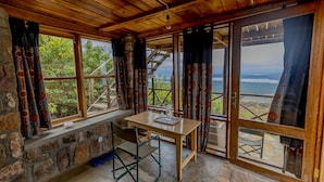 A rustic room with a wooden ceiling, stone walls, a wooden table, and large windows with patterned curtains. - Akagera Rhino Lodge (Kabare)