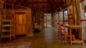 A rustic wooden interior with a thatched roof, stone walls, and a large window offering a view of the outdoors. - Akagera Rhino Lodge (Kabare)