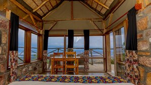 A room with a wooden ceiling, stone walls, a bed, a table with chairs, and large windows offering a view of the sea. - Akagera Rhino Lodge (Kabare)