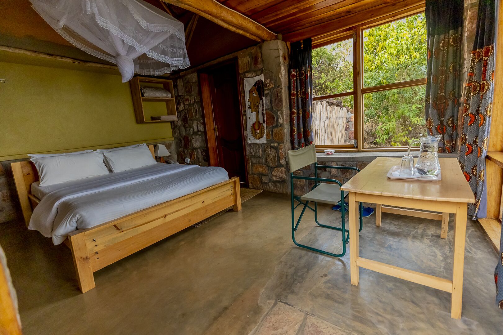 Un lit en bois avec un matelas blanc, une petite table sur laquelle se trouve une carafe, et une chaise dans une pièce rustique avec un mur en pierre et une fenêtre ornée de rideaux.