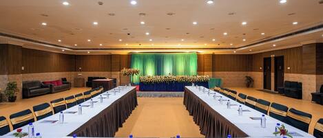 Un salón de conferencias con mesas largas, sillas y un escenario con cortina verde.