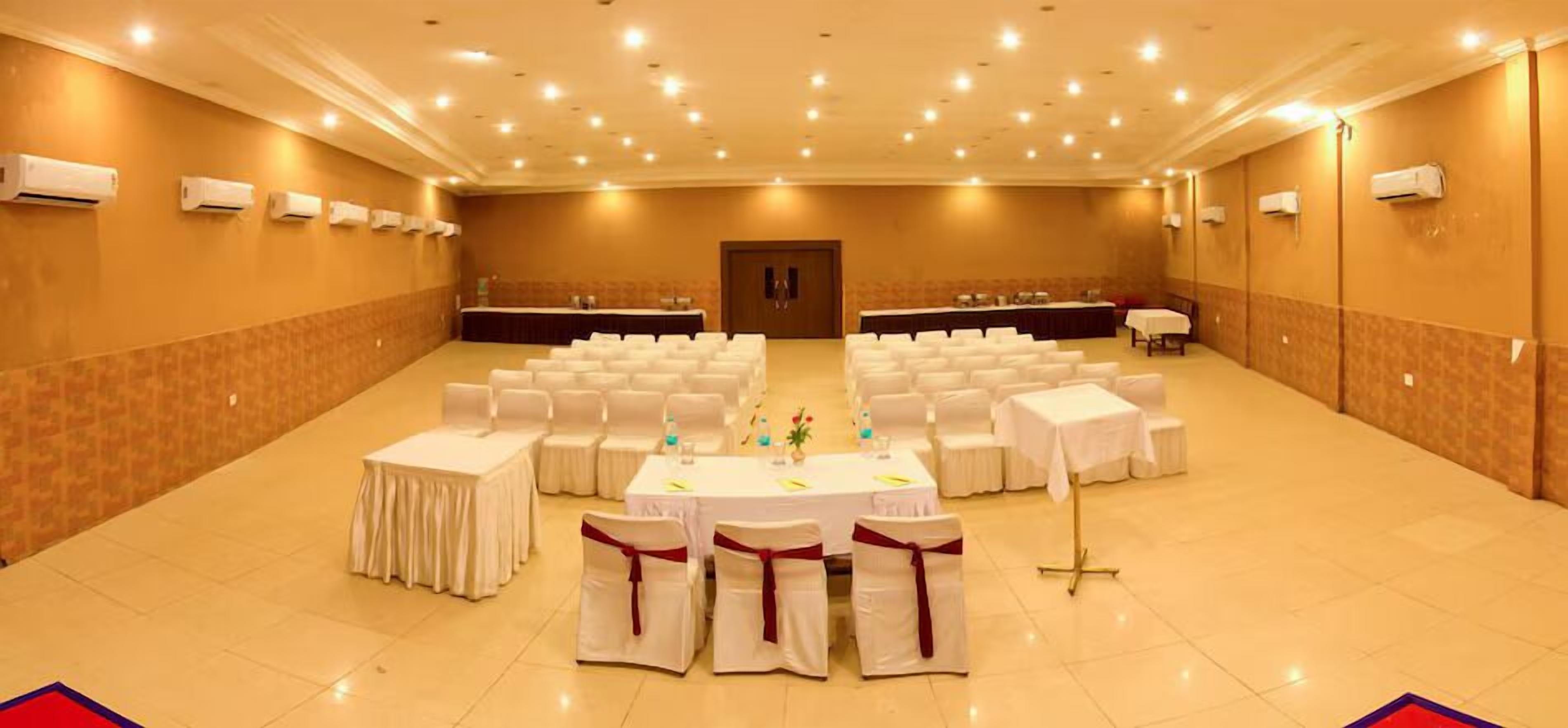 A large conference room with rows of white tables and chairs, air conditioning units on the walls, and a door in the center.