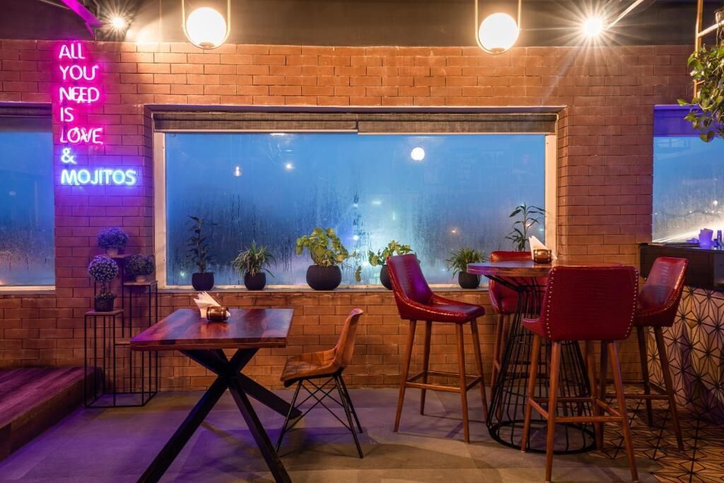 A cozy bar area with brick walls, a neon sign, and a large window overlooking a cityscape.