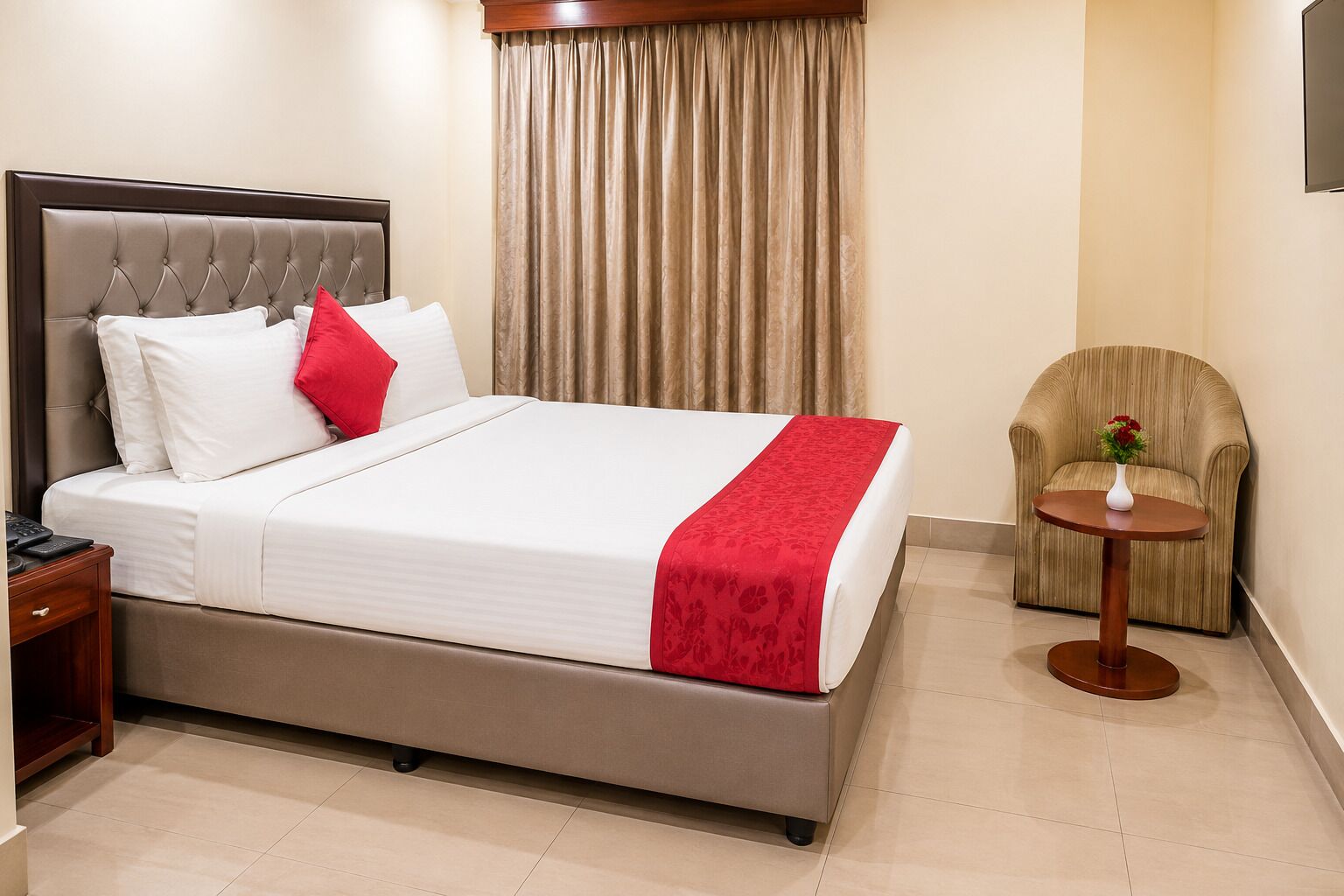 A hotel room with a bed, a nightstand, a chair, and a small table with a vase.