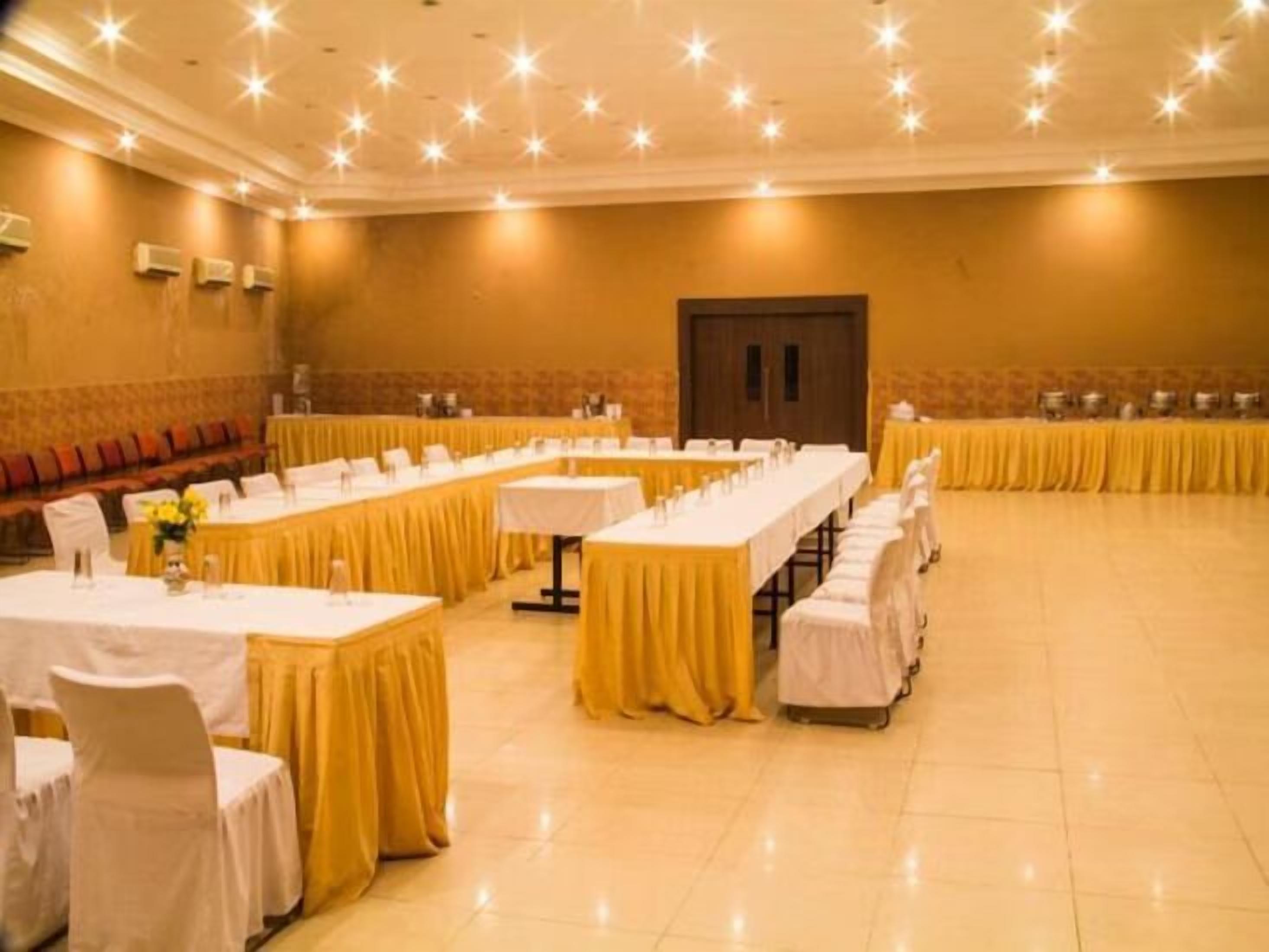 Uma sala de conferências com mesas compridas, cadeiras, palco e uma porta.