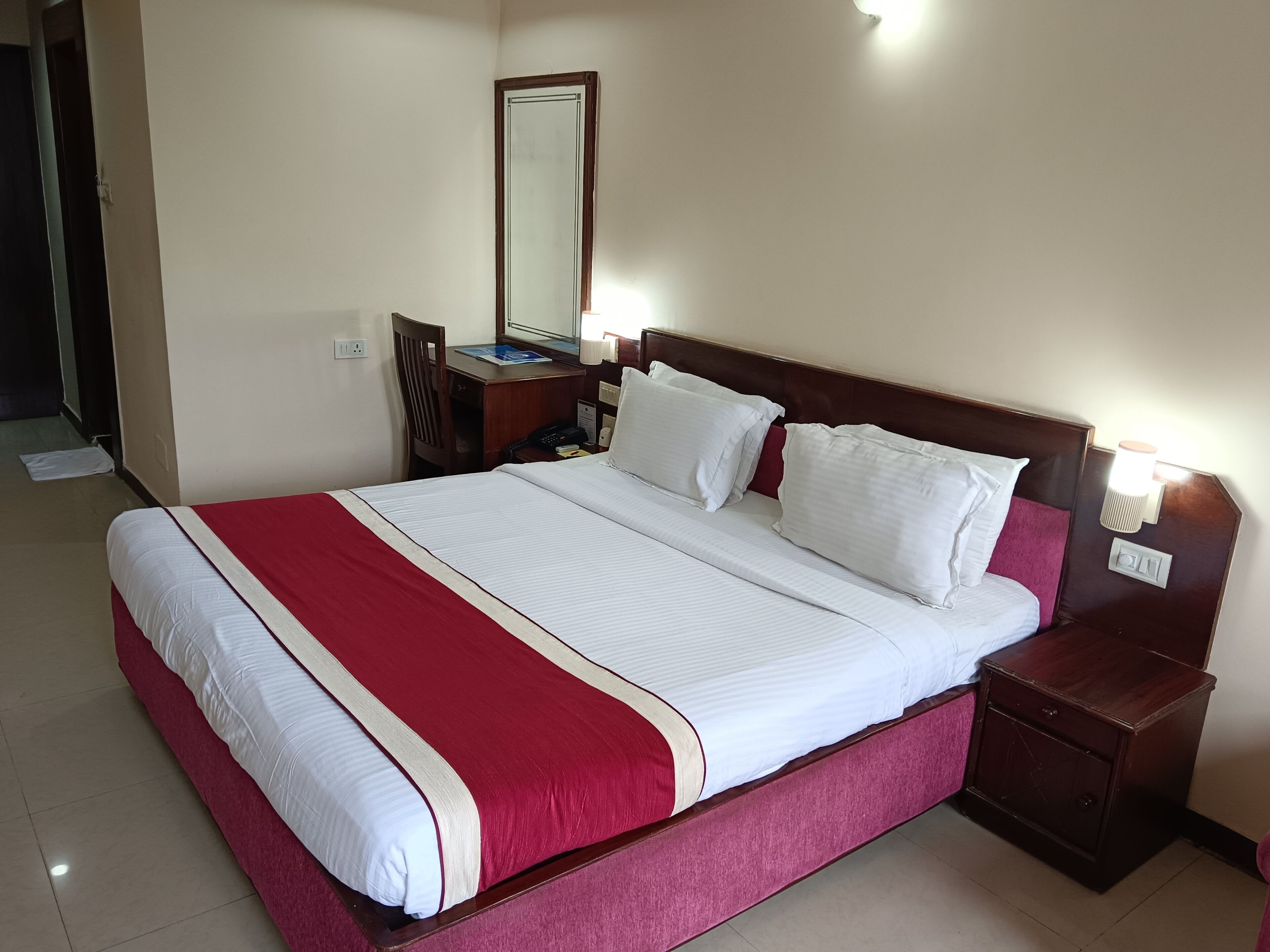 A hotel room with a large bed, a desk, a chair, a nightstand, and a wall-mounted lamp.
