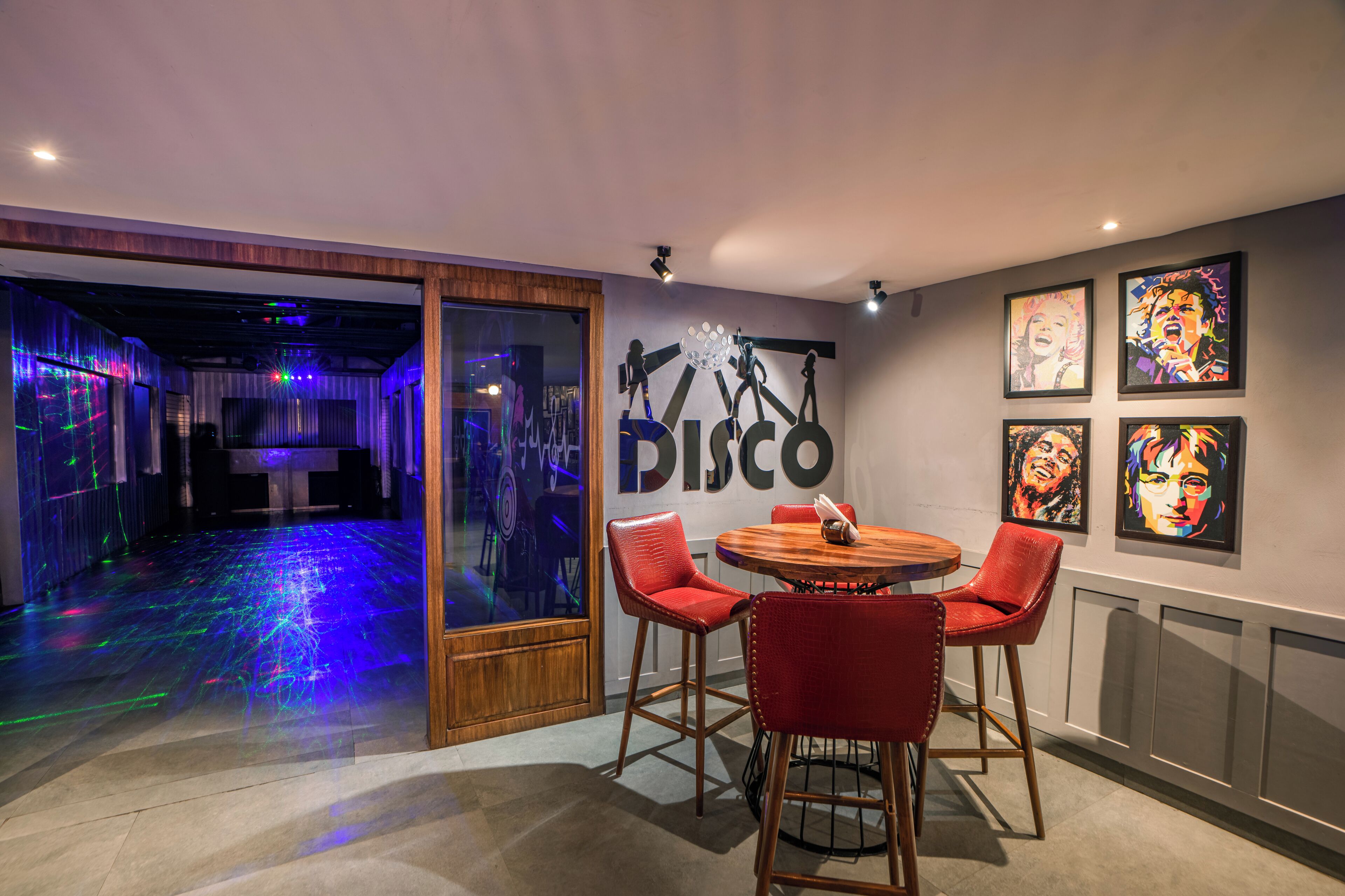 A room with a disco-themed wall art, a round wooden table with red chairs, and a glass door leading to a dance floor with colorful lights.