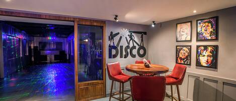 A room with a disco-themed wall art, a round wooden table with red chairs, and a glass door leading to a dance floor with colorful lights.