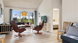 Ett rymligt vardagsrum med ett stort fönster, två bruna läderfåtöljer, ett träbord, en tv på ett stativ och en soffa.