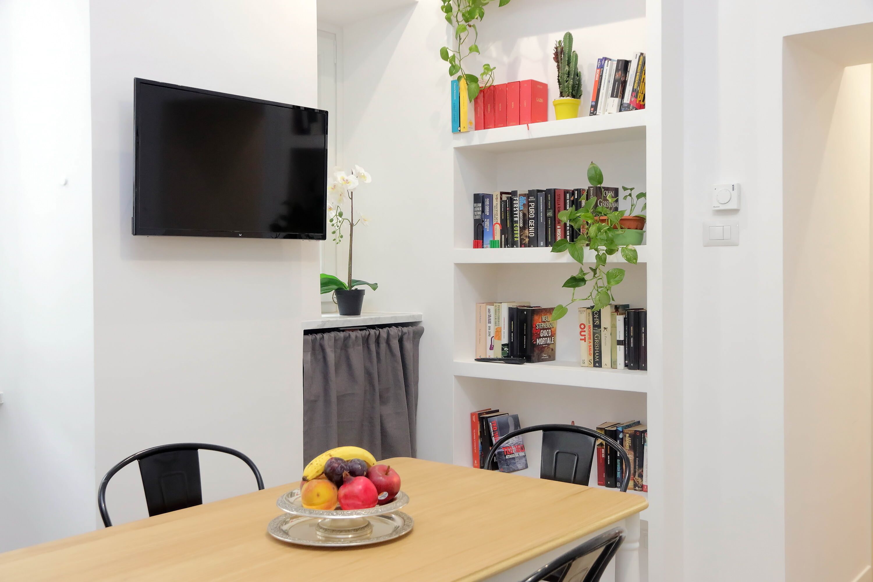 A dining area with a wooden table, chairs, a fruit platter, a bookshelf with books and plants, and a mounted TV.