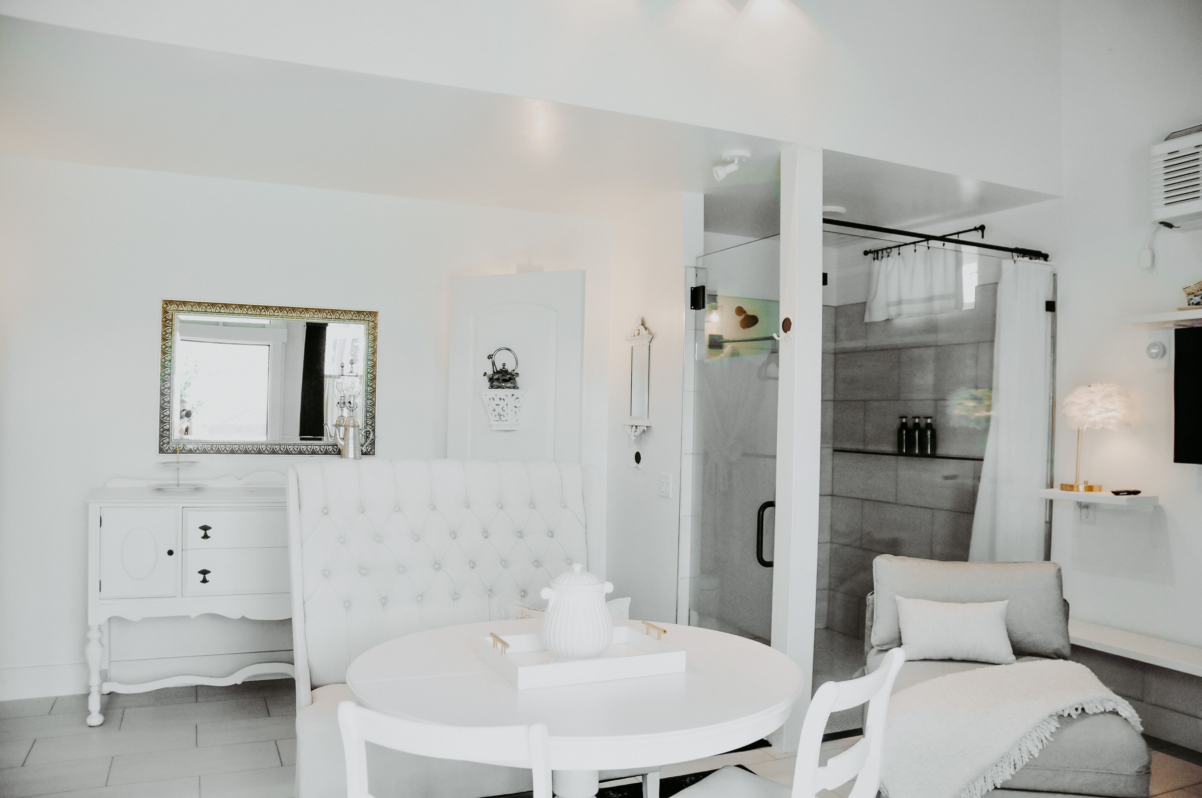 Een moderne, witte kamer met een ronde eettafel, een witte bank, een glazen douchecabine en een witte garderobe met spiegel.