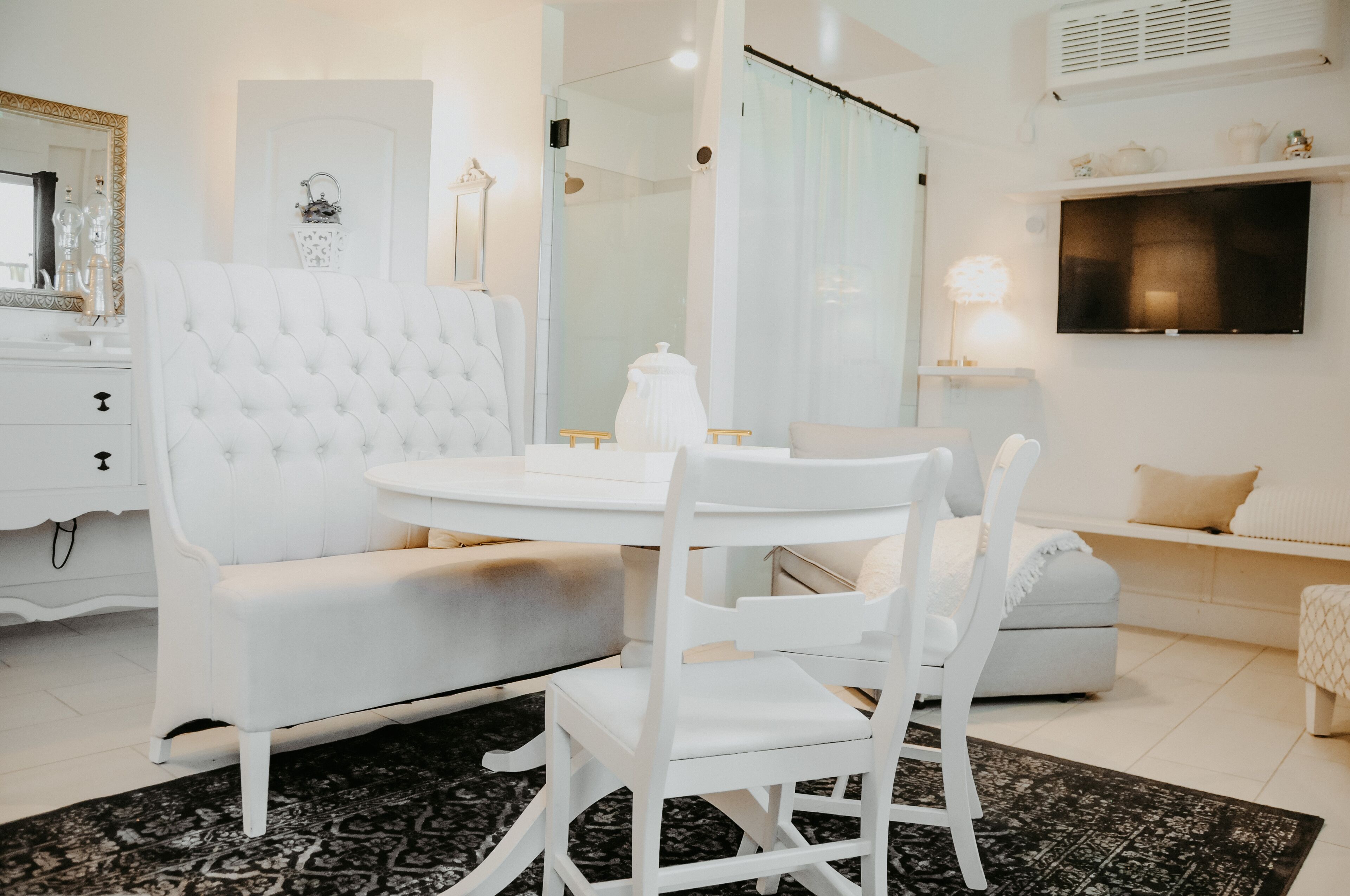 A modern, white-themed bedroom with a round dining table, white upholstered chairs, a white sofa, and a TV mounted on the wall.