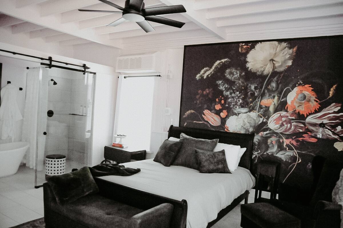 A bedroom with a large bed, a shower, a large floral painting, and a ceiling fan.
