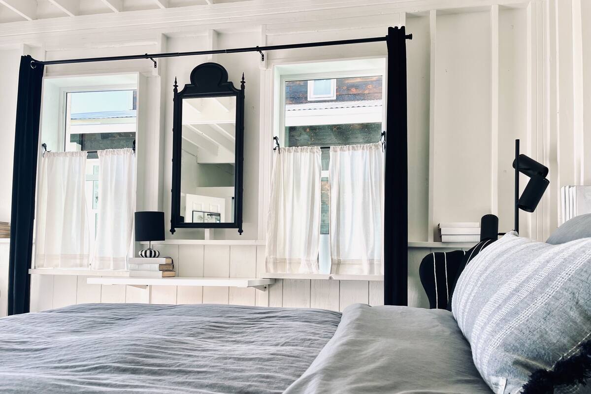 A bedroom with a large bed, a bedside table, a mirror, and a window with curtains.