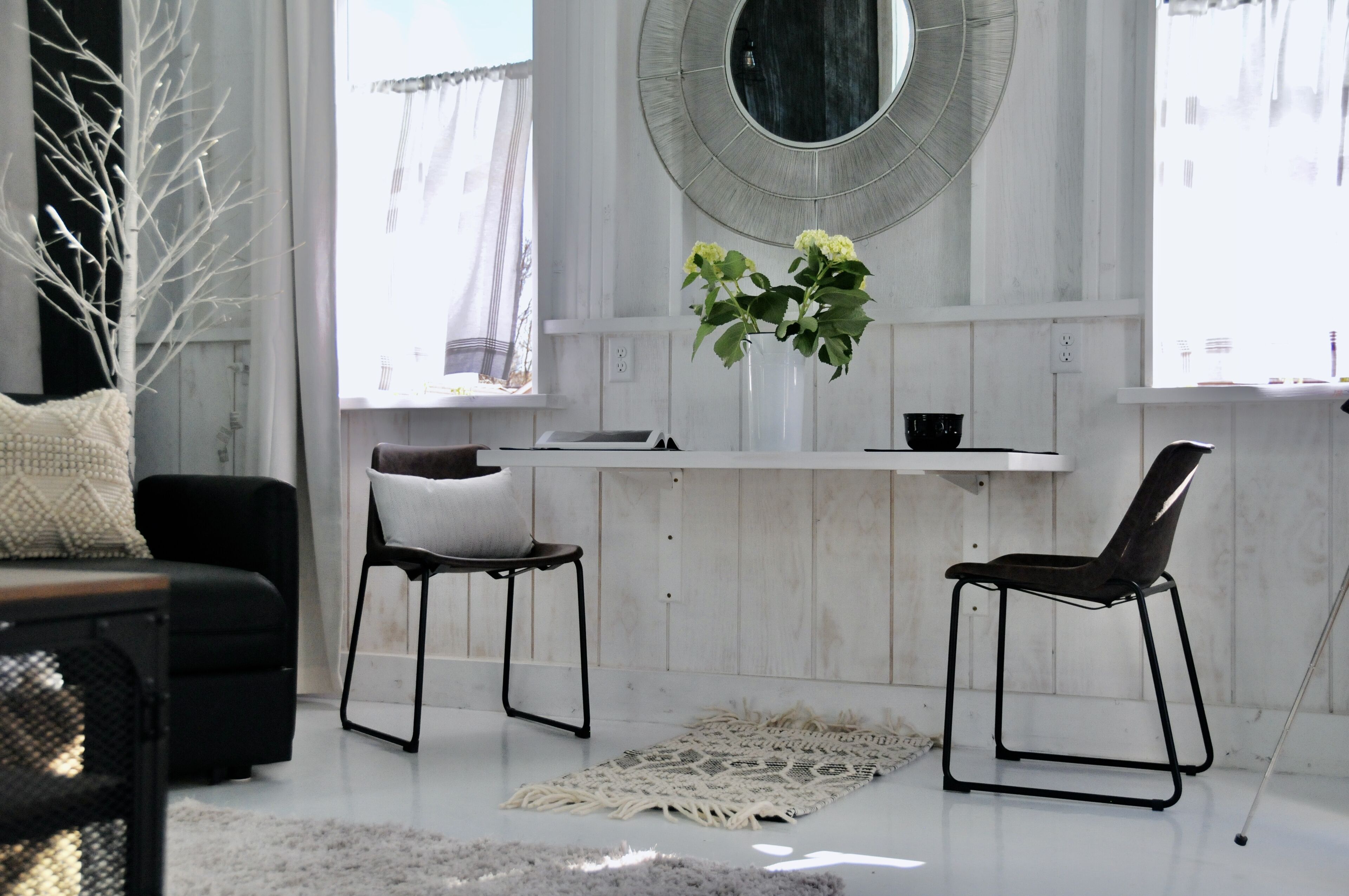 A modern living room with a white wall, a round mirror, a white vase with flowers, two black chairs, a black sofa, and a white rug.