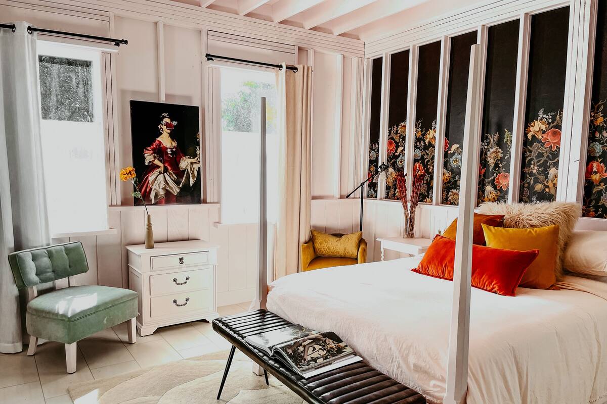 A bedroom with a four-poster bed, a green armchair, a white nightstand, and a large window with floral curtains.