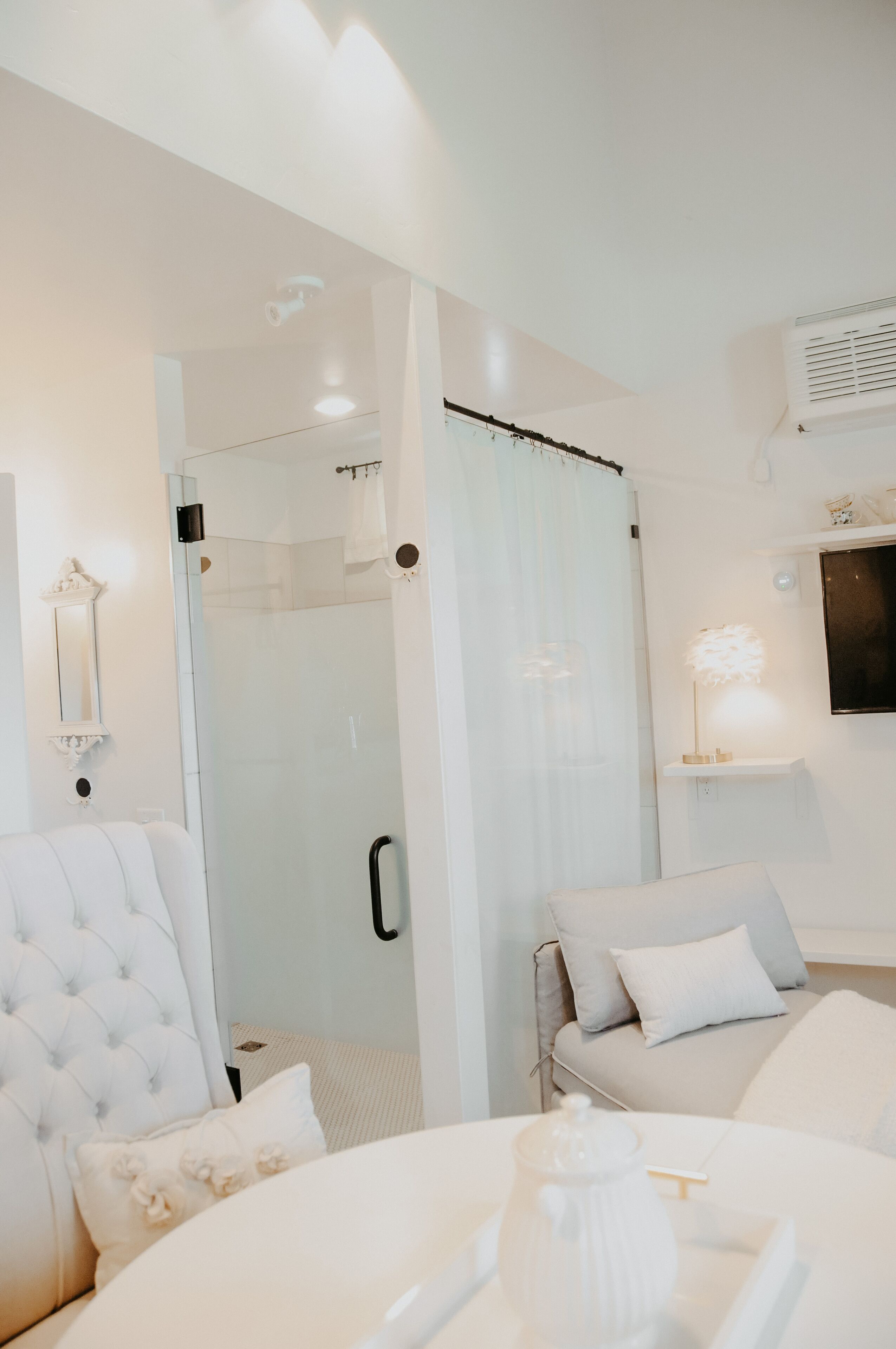 A modern hotel room with a glass shower enclosure, a white bed with a tufted headboard, a white armchair, and a bedside table with a white lamp.