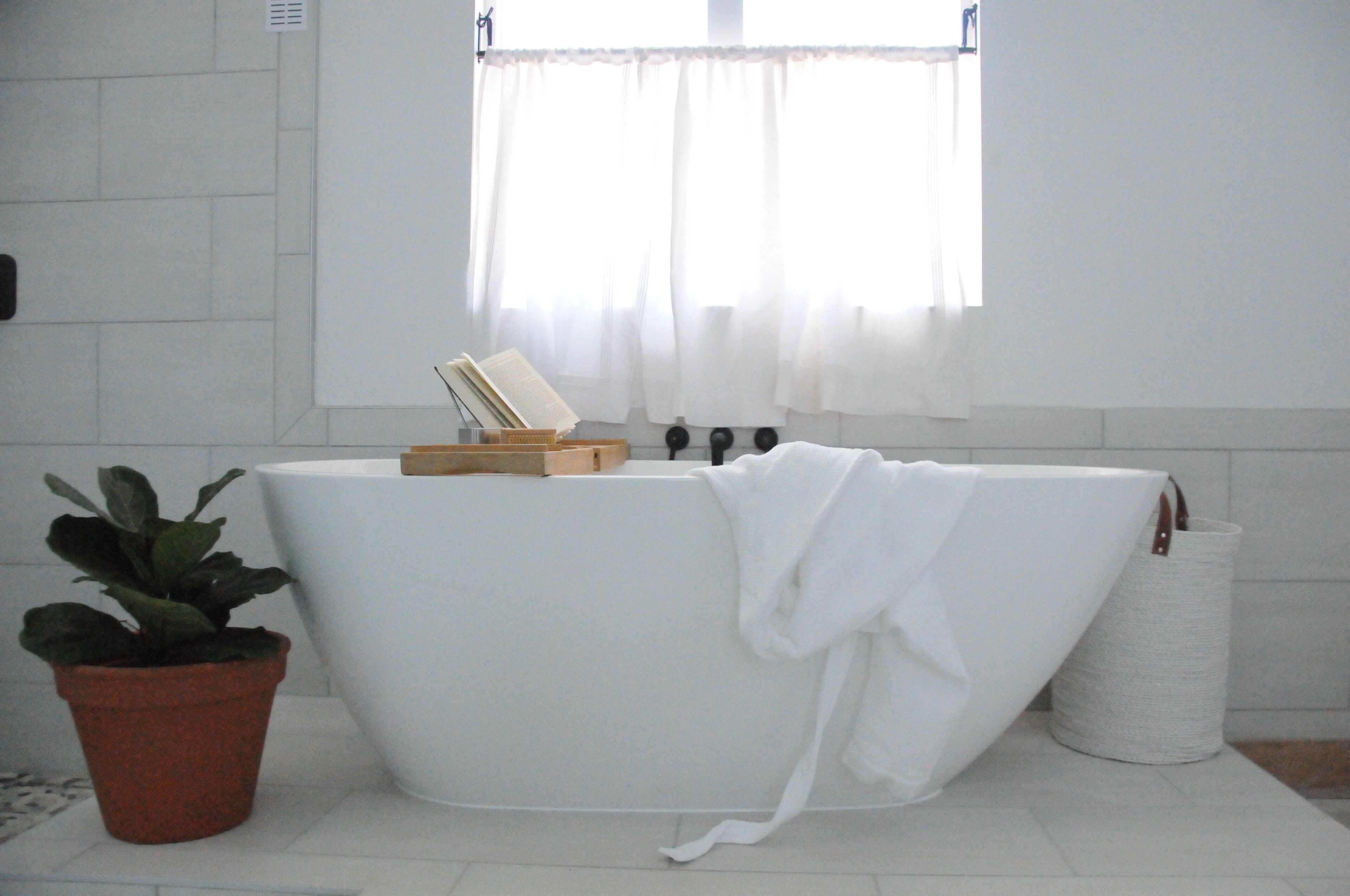 A modern bathroom with a freestanding bathtub, a potted plant, and a window with sheer curtains.