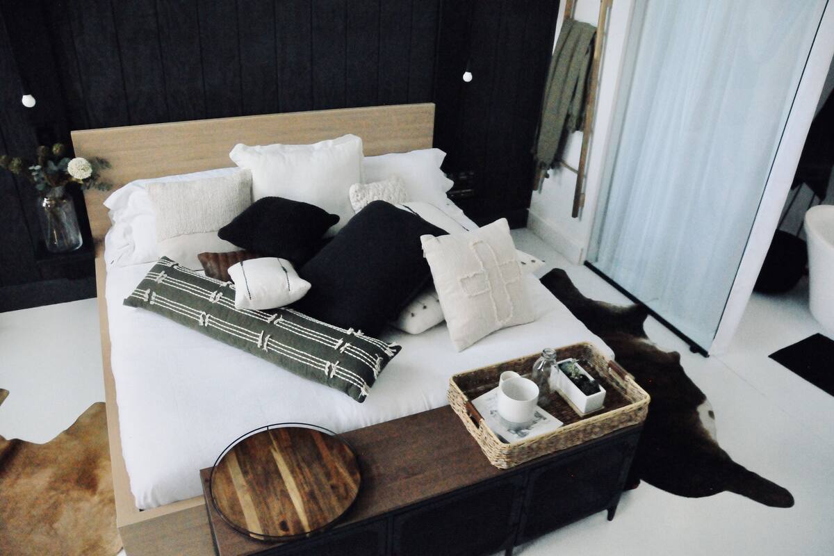 A neatly made bed with multiple pillows and a bedside table with a tray containing a cup and a small plant.