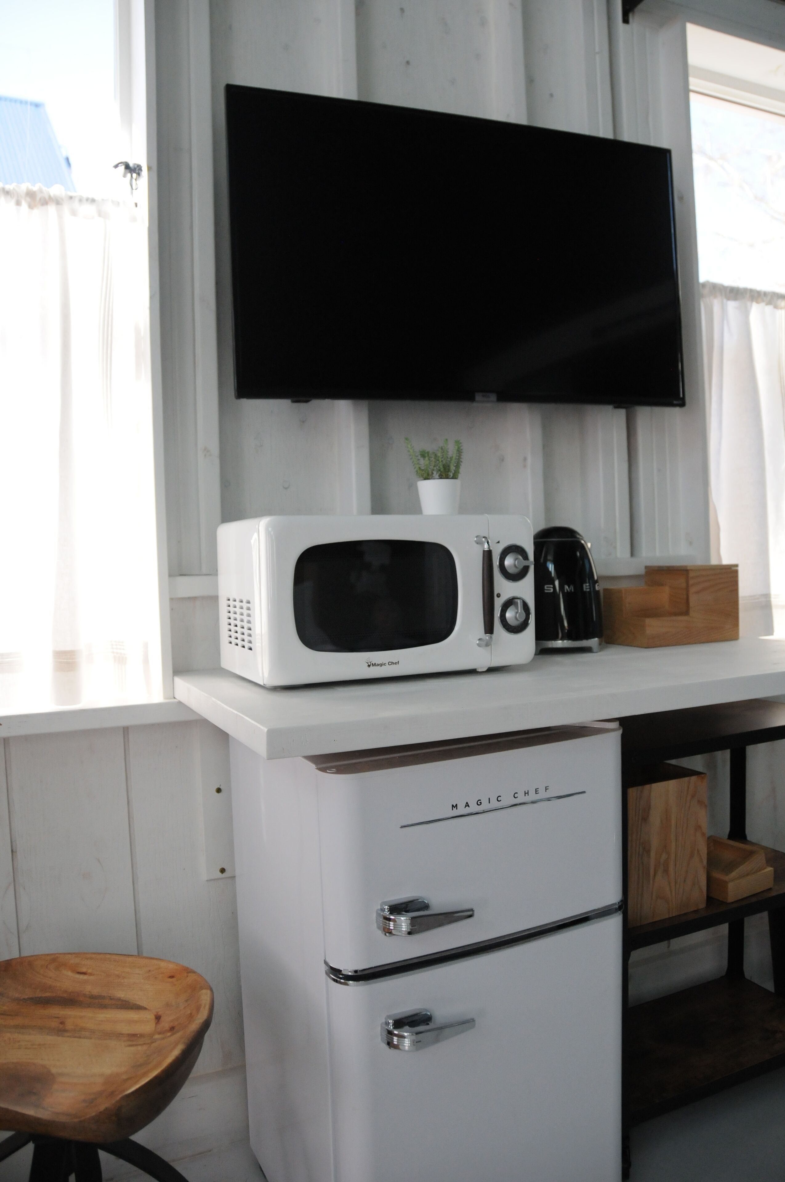 A kitchenette with a white microwave, toaster, and coffee maker on a white countertop. A small potted plant is on the microwave, and a wooden box is on the countertop. There is a white refrigerator with the brand name "MAGIC CHEF" below the countertop.