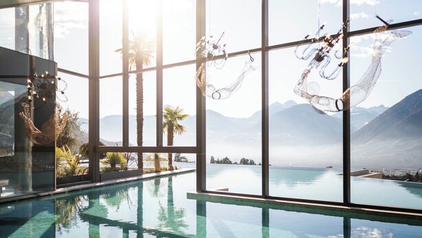 Una piscina a sfioro interna con acqua cristallina, circondata da ampie finestre che offrono vista su montagne e un lago.