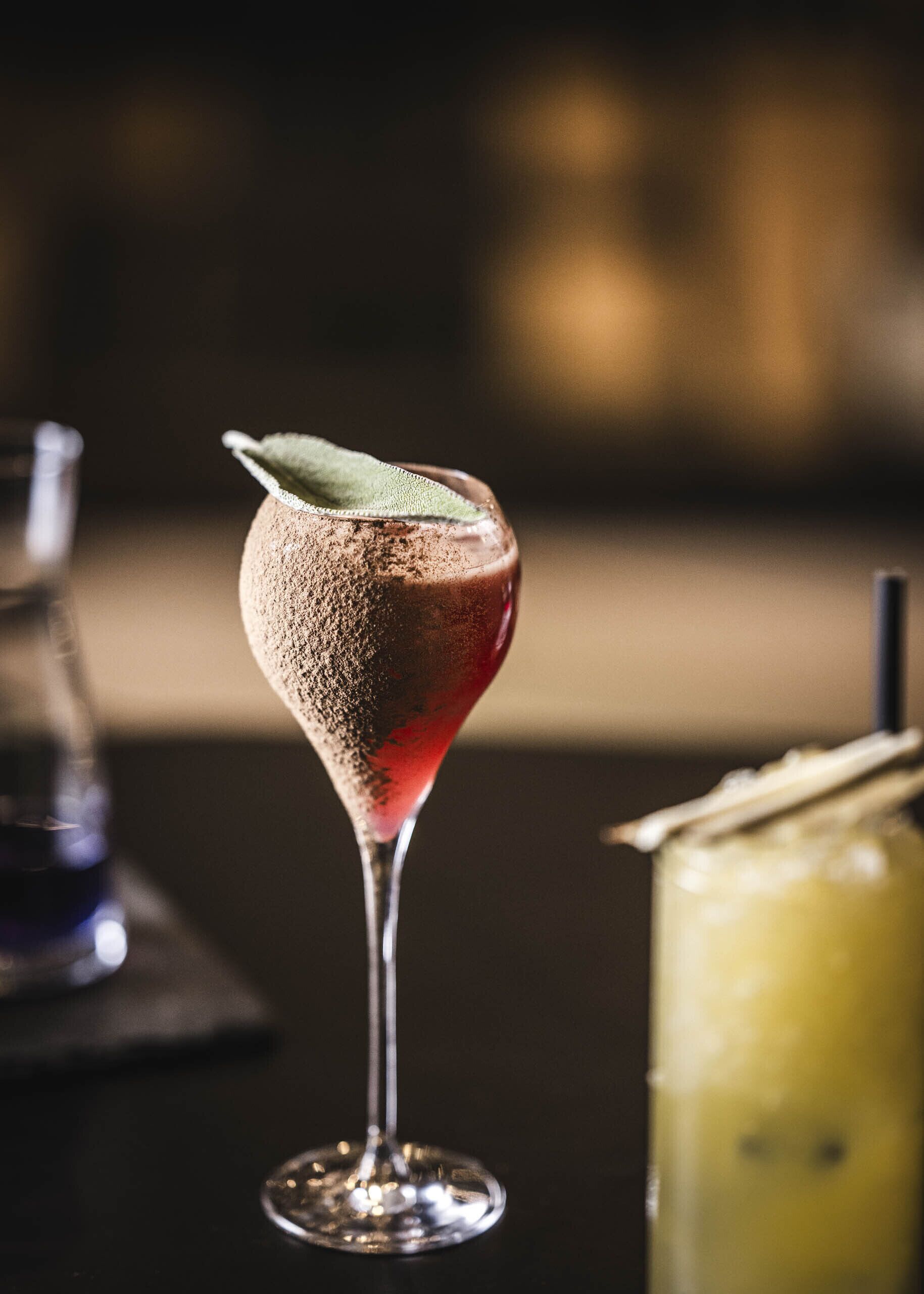 Un cóctel en una copa de martini con una decoración de hoja, una copa de vino tinto y una botella de vino sobre una mesa.