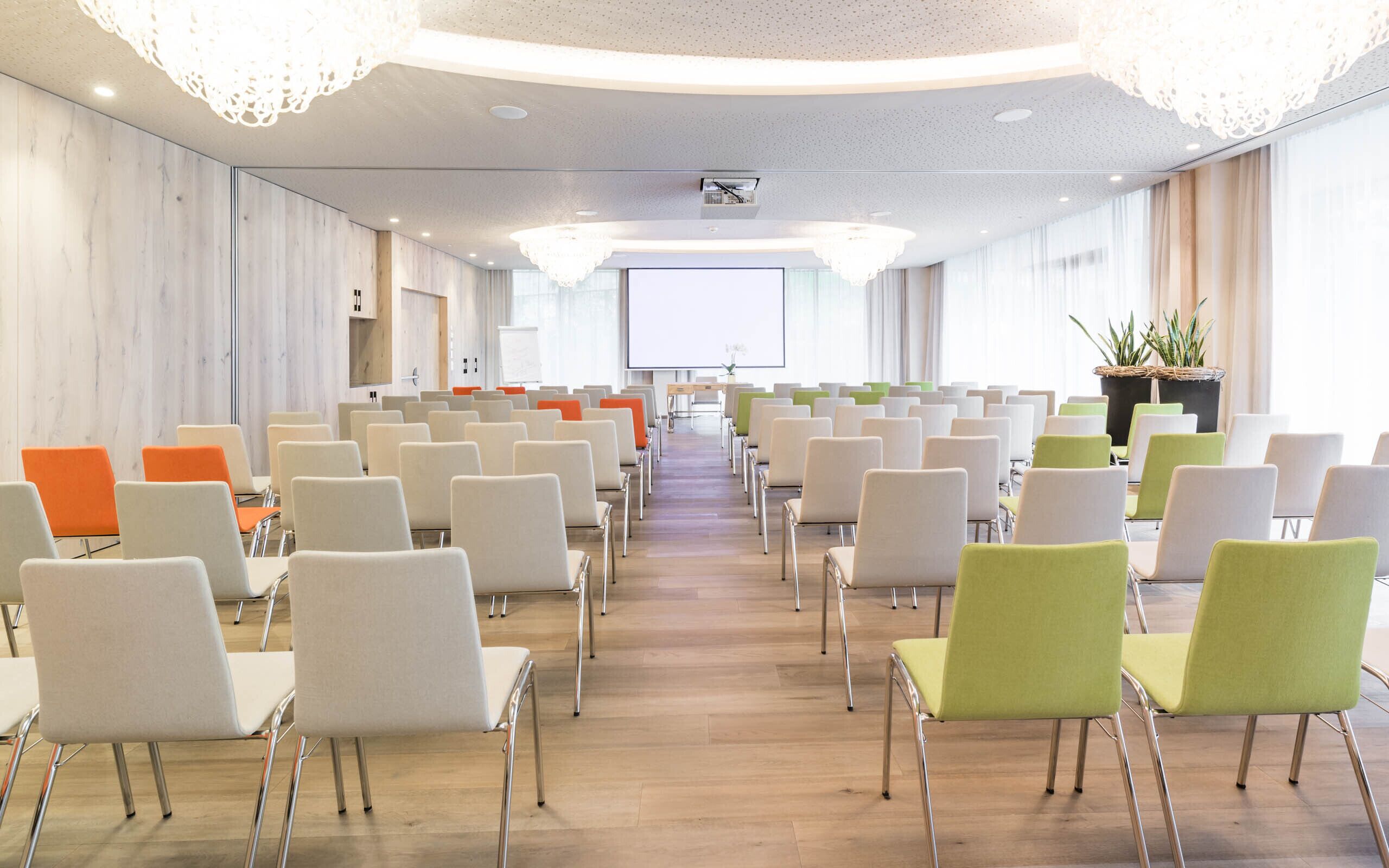 Un salón de conferencias con filas de sillas, una pantalla de proyector y un ventanal grande.