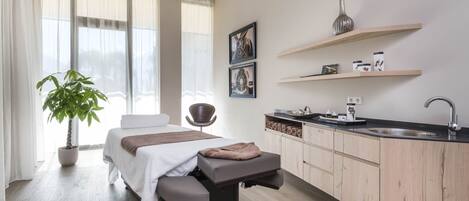 Una habitación moderna con una cama, una camilla de masaje, una planta en maceta, una silla, un lavabo y una pequeña zona de cocina.
