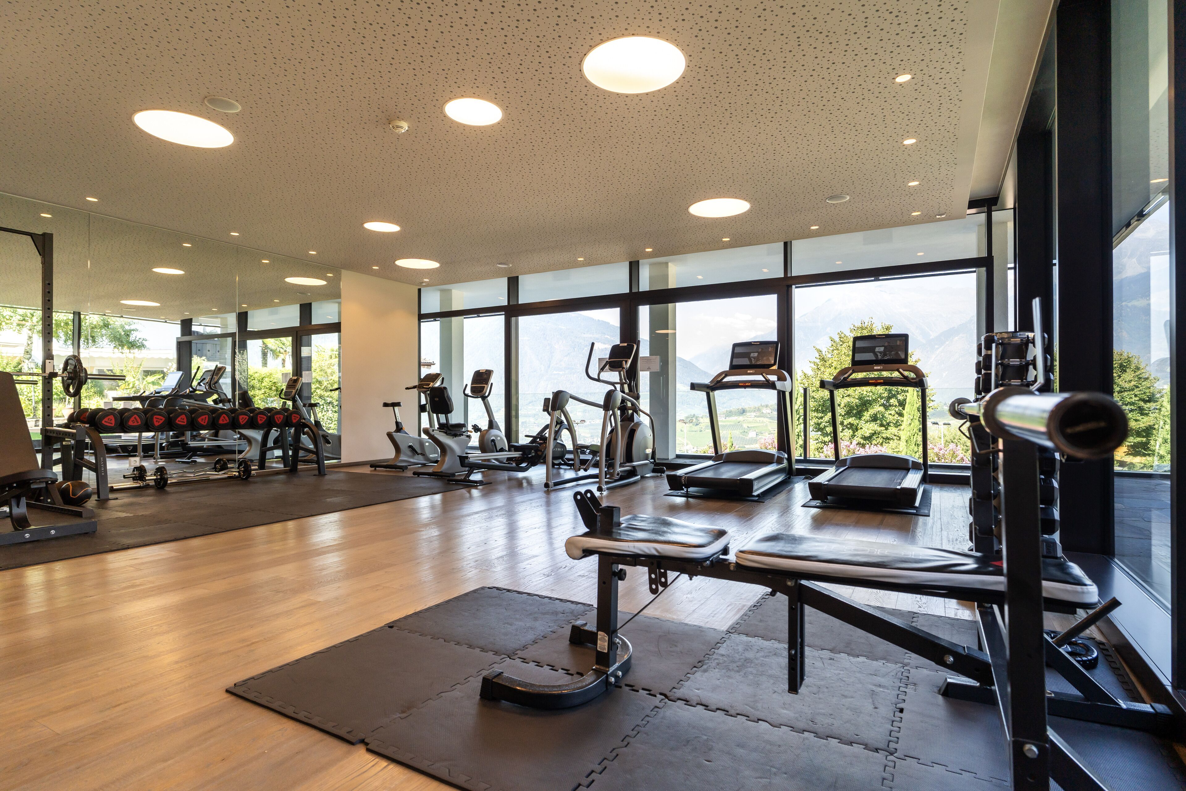 Un gimnasio moderno con diverso equipo de ejercicio, ventanas amplias y suelo de madera.