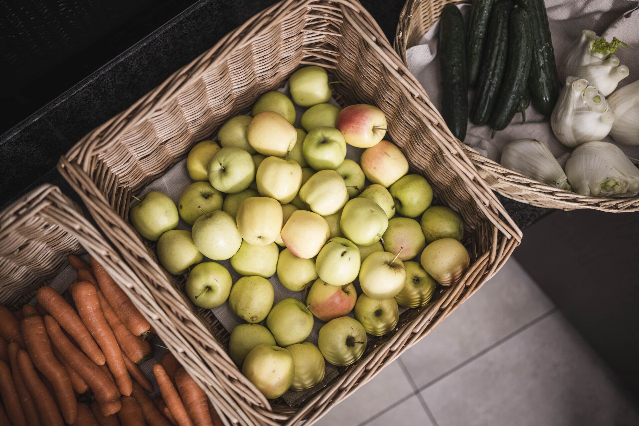Una cesta llena de manzanas verdes, zanahorias, pepinos y cebollas.