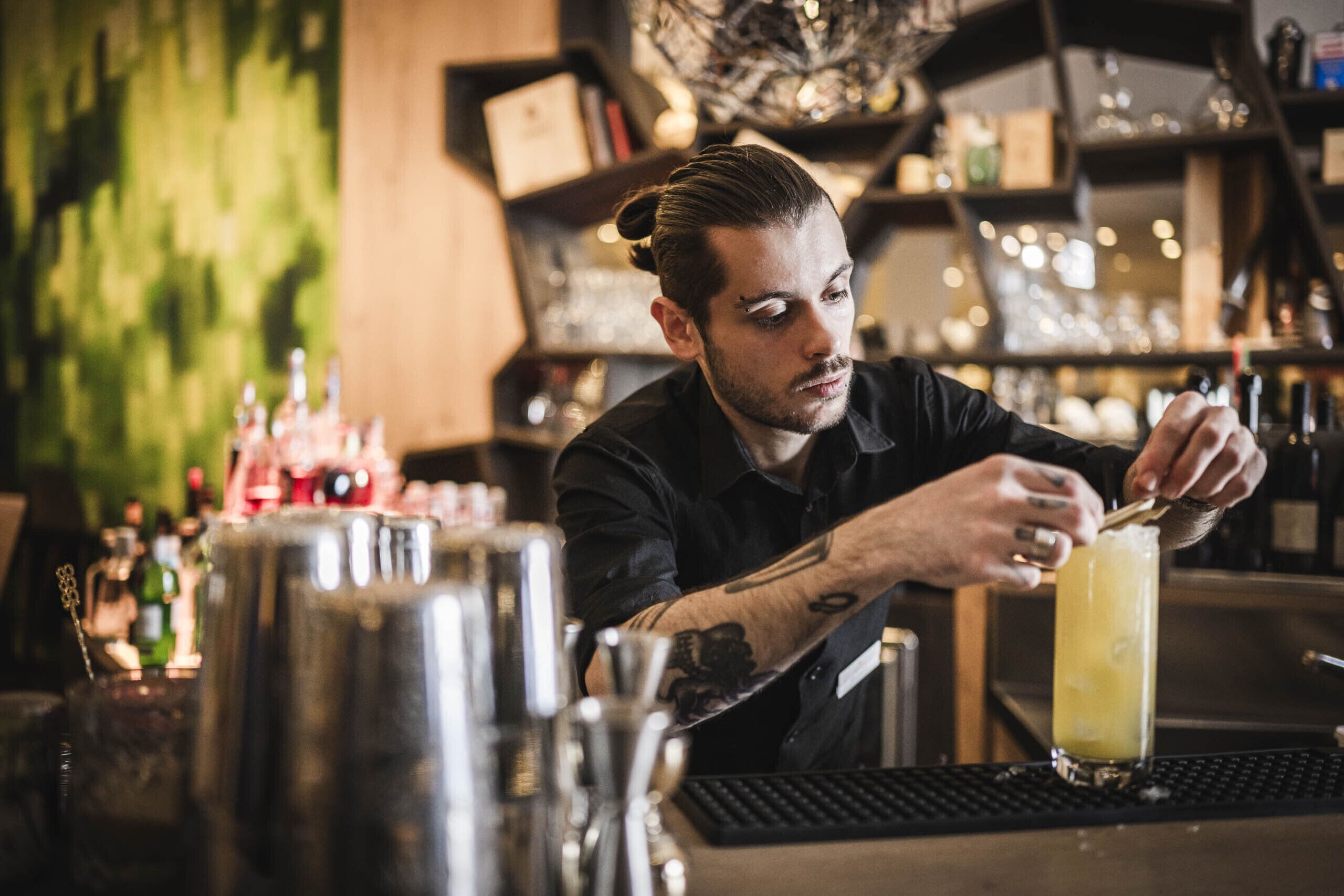 En bartender laver en drink bag en bar med grøn væg og hylder.