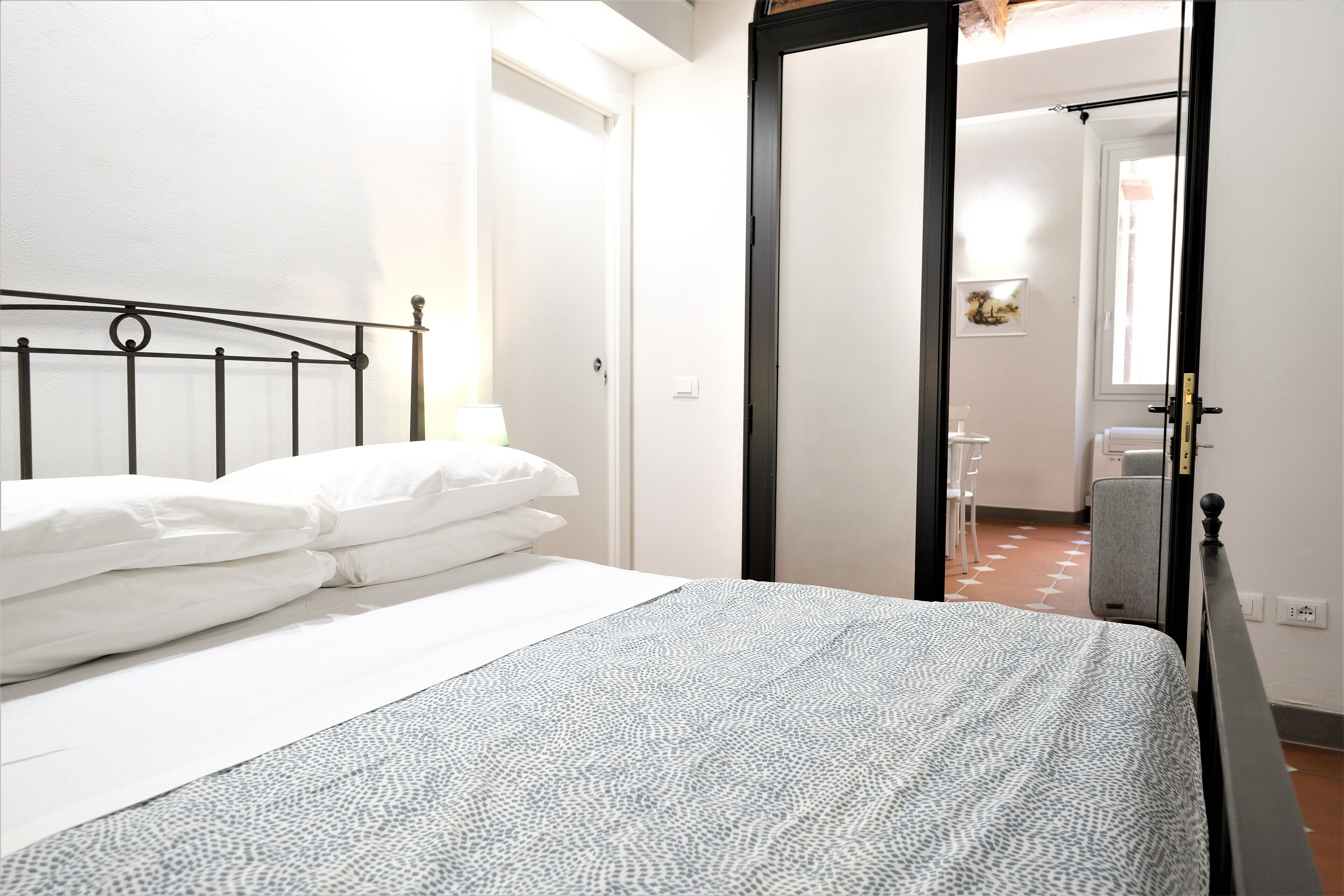 Un lit bien fait, avec du linge de lit blanc et une couvre-lit à motifs, une tête de lit en métal noir et un mur blanc avec une porte donnant sur une autre pièce.