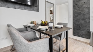 A modern dining area with a rectangular table, grey upholstered chairs, a flat-screen TV mounted on the wall, and a large mirror above the table. - BRIGHT Herzogenaurach Zentrum (Herzogenaurach)