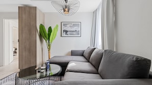 A modern living room with a grey sectional sofa, a glass coffee table, a framed landscape photo, and a large green plant. - BRIGHT Herzogenaurach Zentrum (Herzogenaurach)