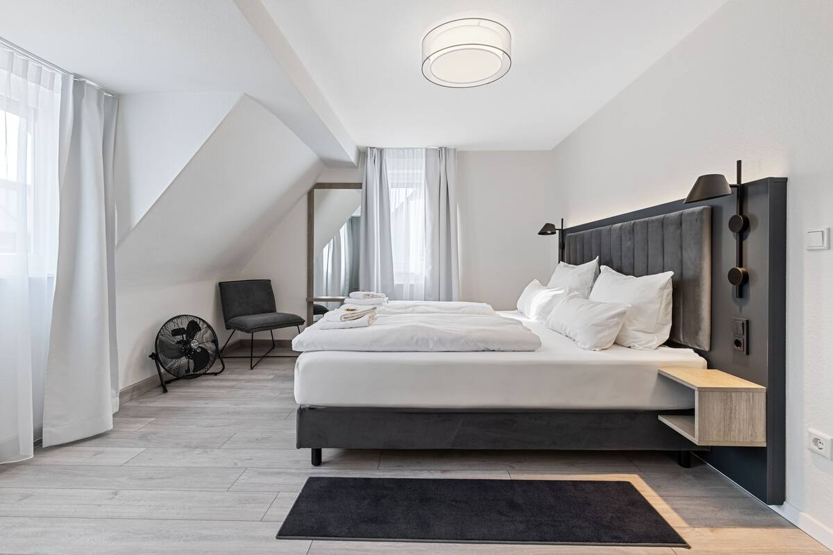 Een moderne slaapkamer met een groot bed, een nachtkastje, een stoel en een ventilator.